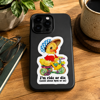 A black phone case featuring a cartoon duck riding a tricycle with the text 'I'm ride or die (until about 9pm or so)', displayed on a wooden desk with a plant, mug, and notebook nearby.