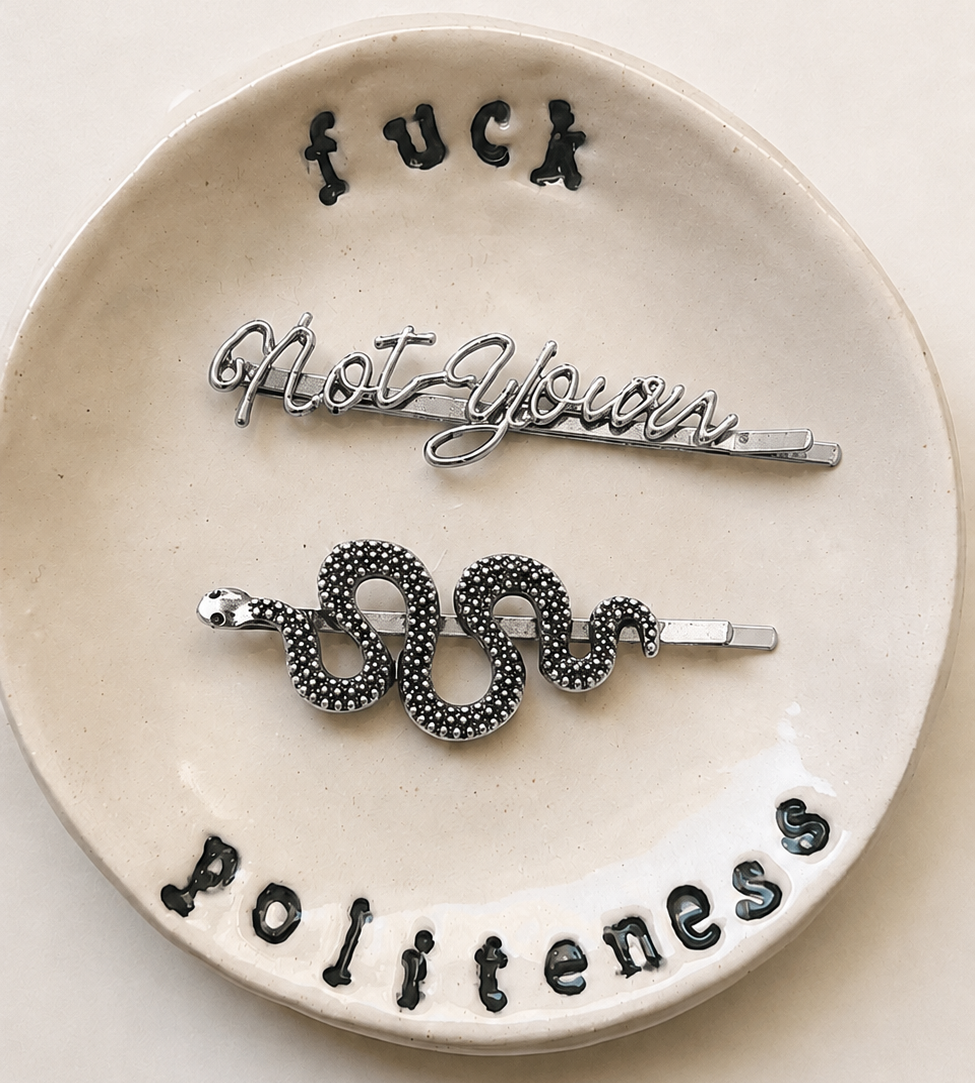 Three metal hair pins displayed on a round beige ceramic dish with black hand-stamped text reading 'fuck', 'not yours', and 'politeness'.