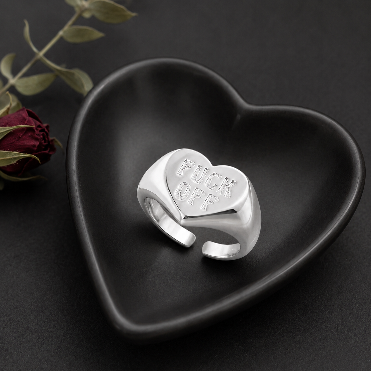 A silver-toned open band ring shaped like a heart, engraved with the words 'FUCK OFF', displayed on a black heart-shaped ceramic dish with a dried rose in the background.