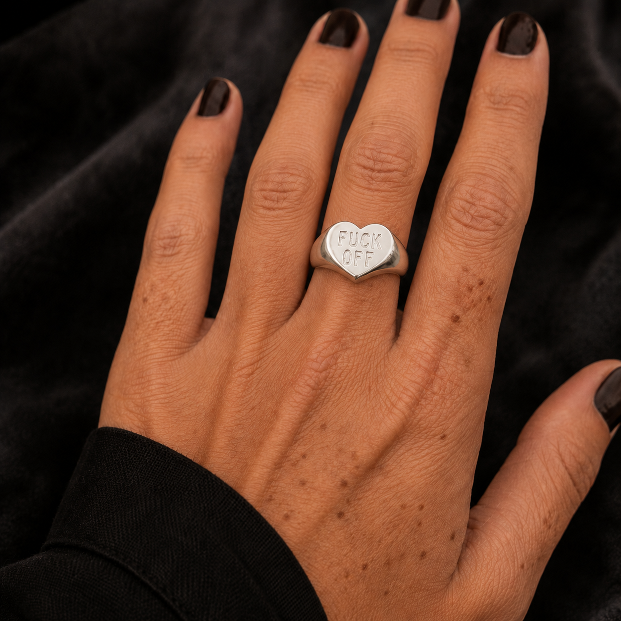 A close-up of a hand wearing a silver heart-shaped ring engraved with the words 'FUCK OFF' in black text, set against a dark textured background.