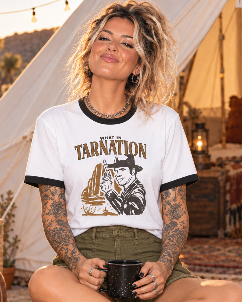 A woman sits cross-legged on a patterned rug in a cozy boho campsite setting, wearing a white ringer t-shirt with black trim and a 'West of Tarnation' graphic print, olive green shorts, and leopard print sneakers.