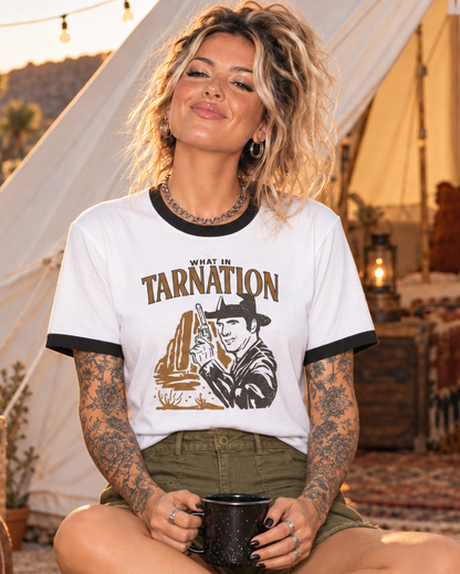 A woman sits cross-legged on a patterned rug in a cozy boho campsite setting, wearing a white ringer t-shirt with black trim and a 'West of Tarnation' graphic print, olive green shorts, and leopard print sneakers.