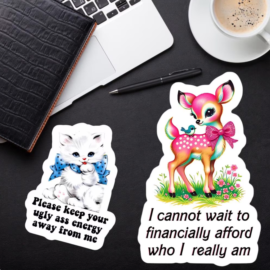 Two die-cut stickers displayed on a dark desk with a laptop, notebook, pen, and coffee cup. One sticker shows a white kitten with a blue bow tie and the text 'Please keep your ugly ass energy away from me'.