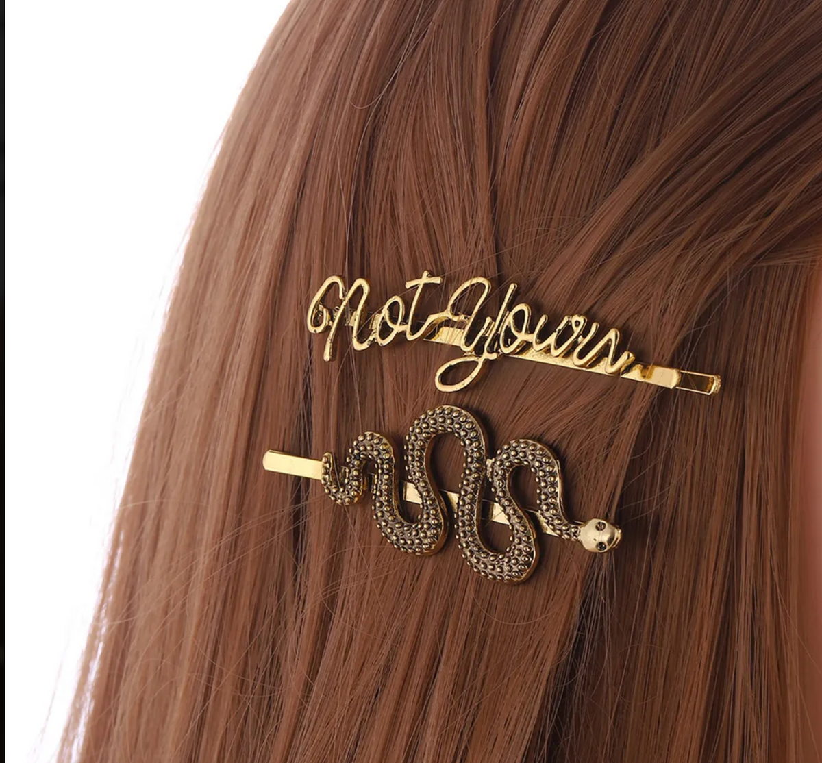 Two gold hair accessories styled on brown hair: a script letter hair clip reading 'not yours' and a snake-shaped hair claw clip with rhinestone detailing.