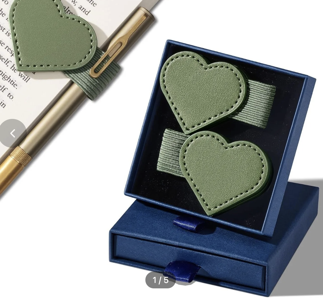 Two olive green heart-shaped leather accessories with stitched edges and elastic bands, displayed inside an open dark blue gift box. A matching pen and a book page are partially visible in the upper left on a white background.