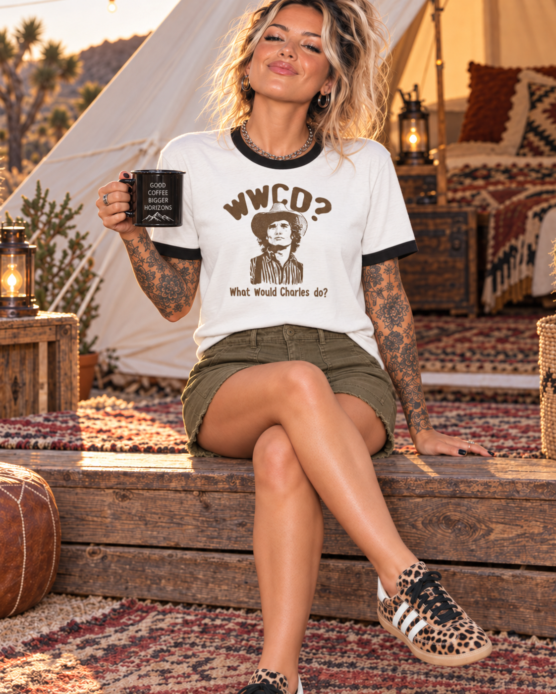 A woman sits on a wooden bench in a rustic outdoor setting, wearing a white ringer T-shirt with black trim and a graphic print that reads 'WWCD? What Would Charles do?'. She is also wearing olive green shorts and leopard print sneakers, holding a black mug.