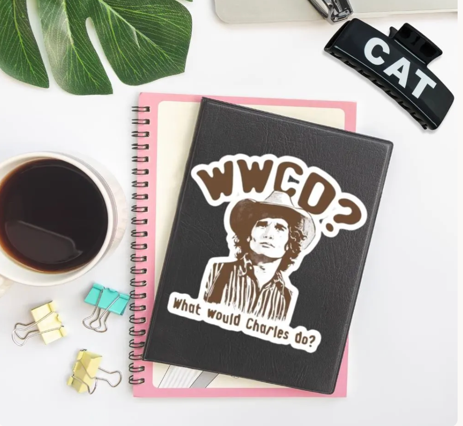 A black spiral notebook with a die-cut sticker featuring the text 'WWGO? What would Charles do?' and a portrait illustration, styled on a white desk with a pink page insert, coffee cup, binder clips, and a leaf.