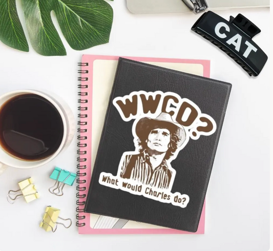 A black spiral notebook with a die-cut sticker featuring the text 'WWGO? What would Charles do?' and a portrait illustration, styled on a white desk with a pink page insert, coffee cup, binder clips, and a leaf.