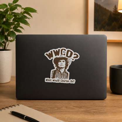 A black laptop on a wooden desk with a die-cut sticker of a man in a cowboy hat and the text 'WWCEO? What would Charles do?' on the back. The scene is styled with a potted plant, a notebook, and a mug in a warm indoor setting.