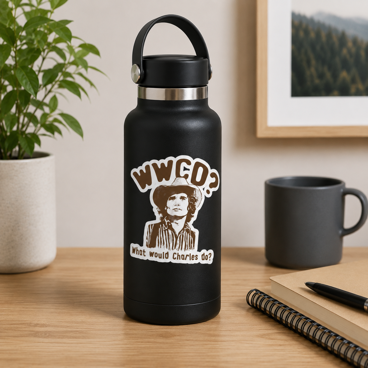 A matte black insulated water bottle with a carry loop stands on a wooden desk. The bottle has a brown and cream graphic print featuring the text 'WWCD? What would Charles do?' and an illustrated portrait.