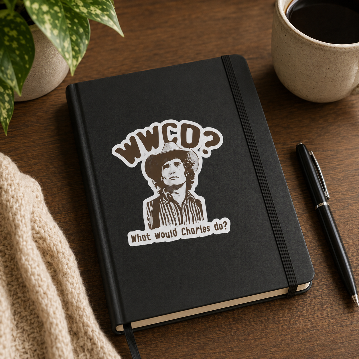 A black hardcover notebook with an elastic closure band, featuring a die-cut graphic of a man in a hat with the text 'WWCD? What would Charles do?'. The notebook is styled on a wooden desk next to a mug of coffee, a black pen, a knit sweater, and a potted plant.