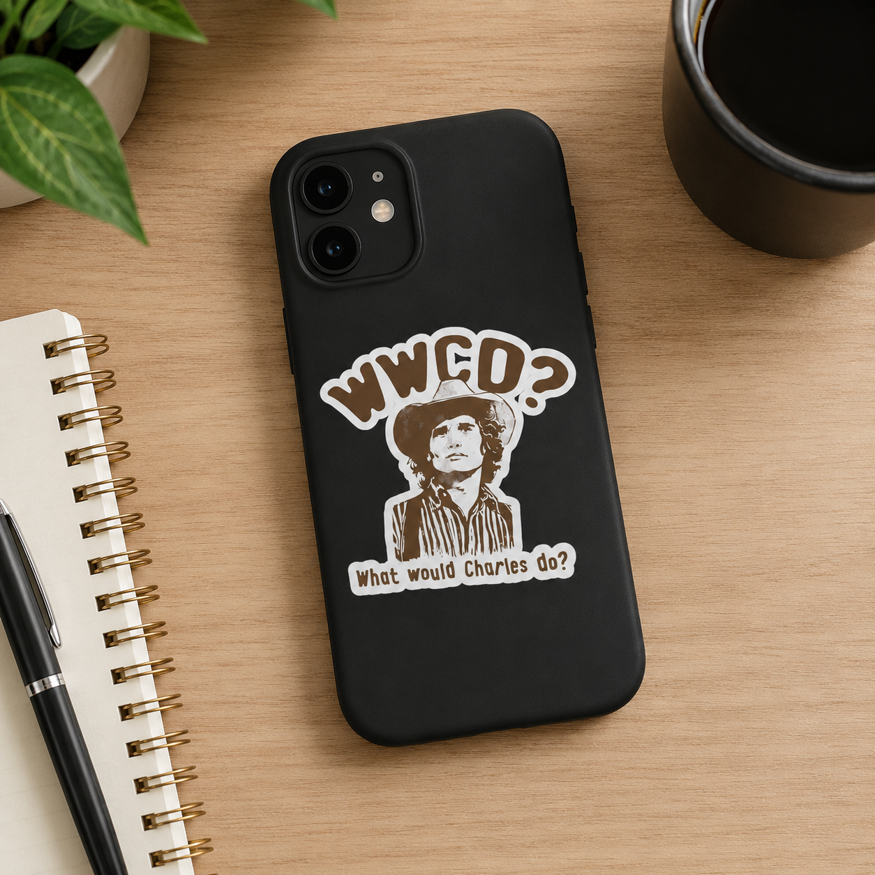 A black phone case featuring a vintage-style graphic of Charles Manson with the text 'WWCD? What would Charles do?' displayed on a wooden desk alongside a notebook, pen, and coffee cup.