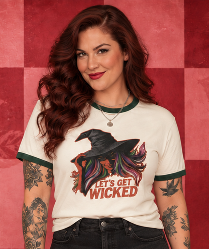 A woman stands against a red patterned checkered wall wearing a white ringer T-shirt with a black graphic and the words 'IT'S NOT WICKED', black denim shorts, and black ankle boots.