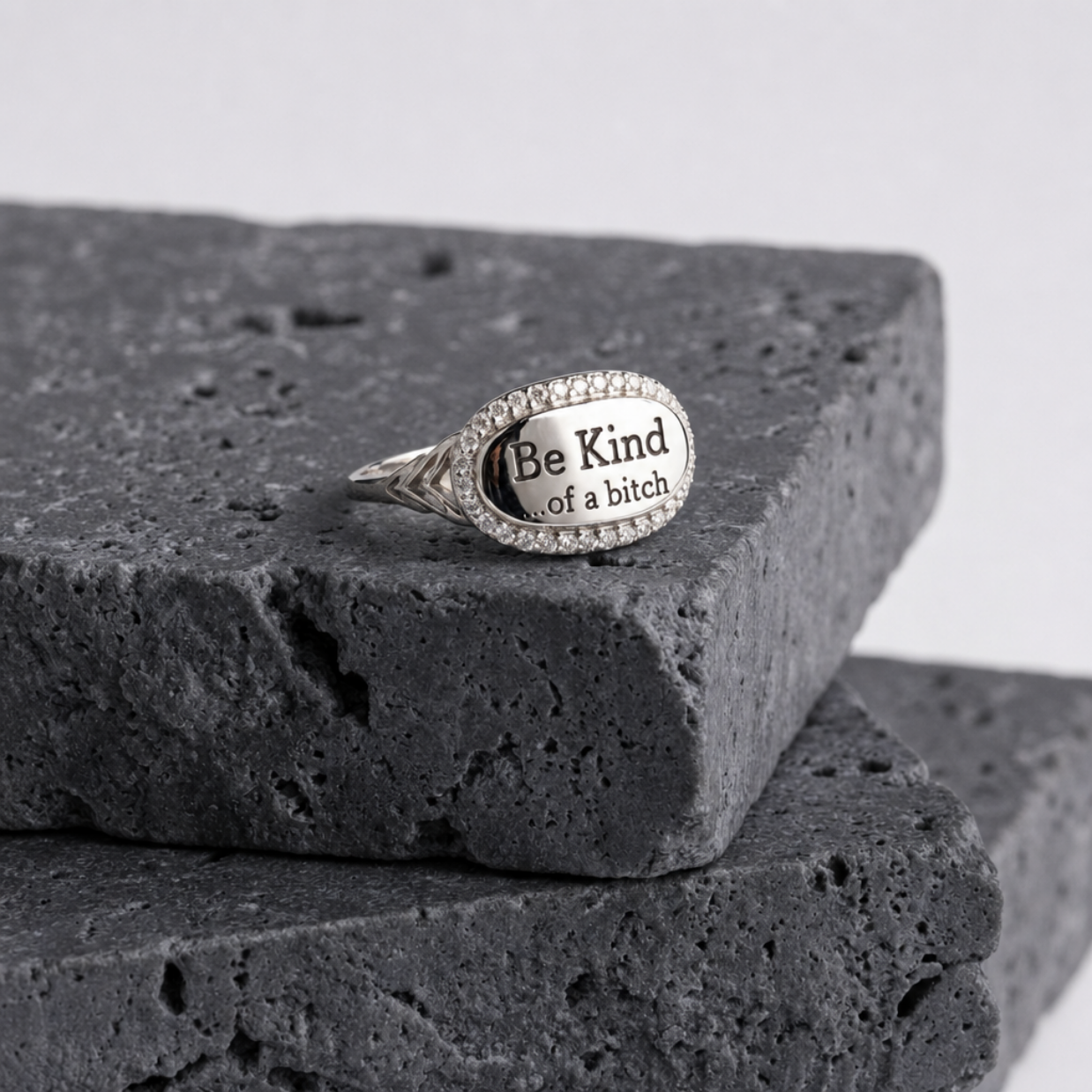 A silver oval signet ring with the text 'Be Kind of a bitch' engraved on the face, resting on a dark textured stone block against a light gray background.
