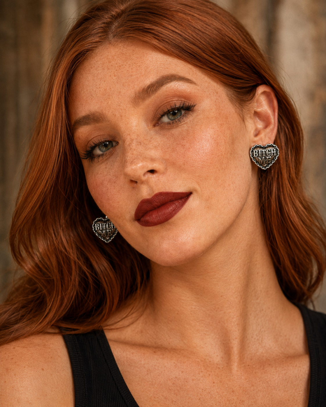 Close-up portrait of a woman with red hair wearing heart-shaped silver earrings with black lettering that reads 'BLOOM'.