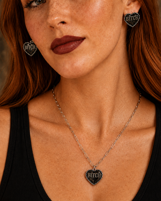 Close-up of a woman wearing a silver chain necklace with a heart-shaped pendant engraved with 'BITCH' in a gothic font on a black textured background. She is also wearing matching heart-shaped stud earrings.