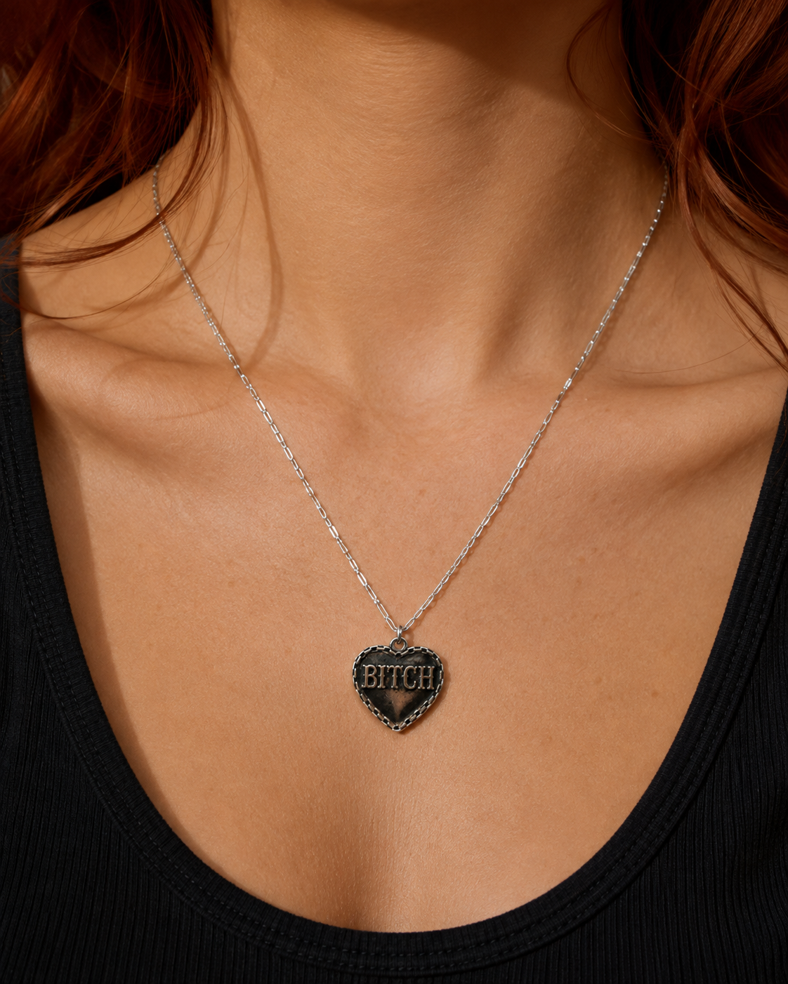 A close-up of a model wearing a silver chain necklace with a heart-shaped pendant that reads 'BITCH' in gold lettering on a black background.