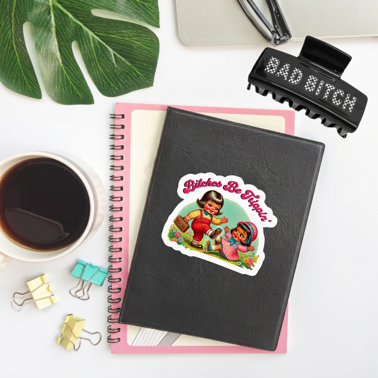 A black spiral notebook with a die-cut sticker that reads 'Bitches Be Hippies' featuring a retro illustration of two people outdoors, styled on a white desk with a pink page, coffee cup, binder clips, a leaf, and a black hair claw clip.