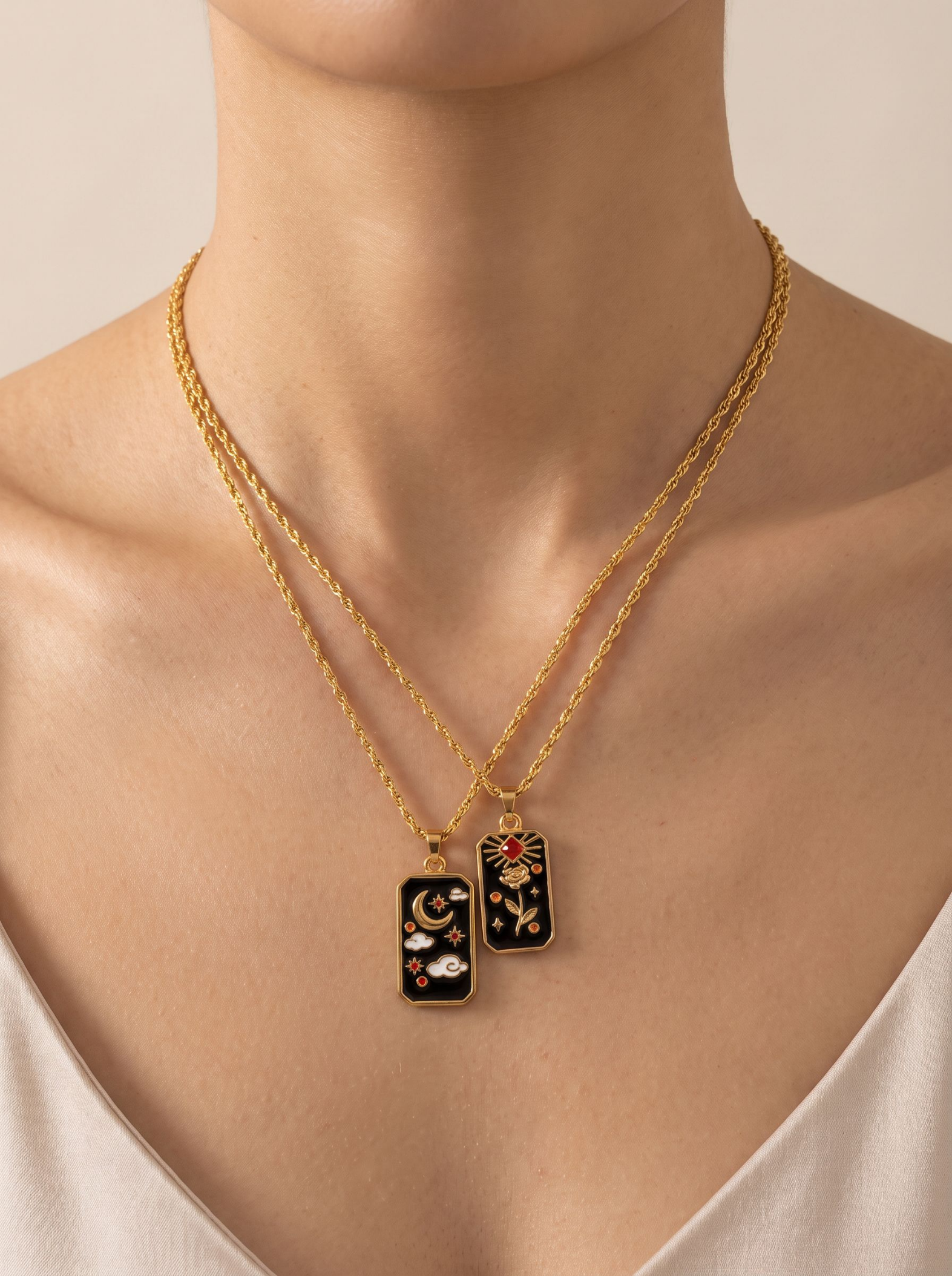 A close-up of a model's neck and chest wearing two layered gold necklaces with rectangular black enamel pendants. One pendant features a gold crescent moon, clouds, and stars, while the other features a gold tiger head with red and white details.