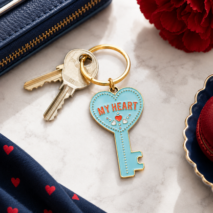 A gold-tone keyring with two keys and a light blue enamel key-shaped charm that reads 'MY HEART' in red lettering, styled on a marble surface with navy fabric, a red flower, and a zipper pouch nearby.