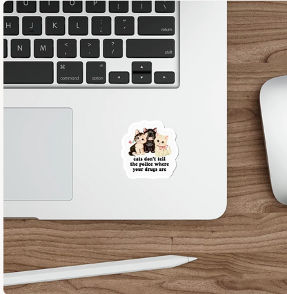 A die-cut sticker featuring three cartoon cats and the text 'cats don't tell the police where your drugs are' is shown on the lower right corner of a silver laptop, placed on a wooden desk next to a white stylus and mouse.