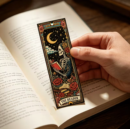 A hand holds a decorative illustrated bookmark shaped like a book cover featuring a skeleton, crescent moon, stars, roses, and the text 'THE READER' over an open book on a dark wooden surface.