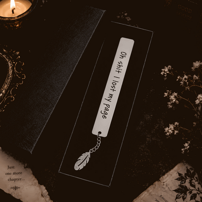 A silver metal bookmark with a feather charm lies on a dark book next to its black cover. The bookmark has handwritten text reading 'Oh sh*t I lost my page' and is styled with a candle, old book pages, and dried flowers on a dark background.