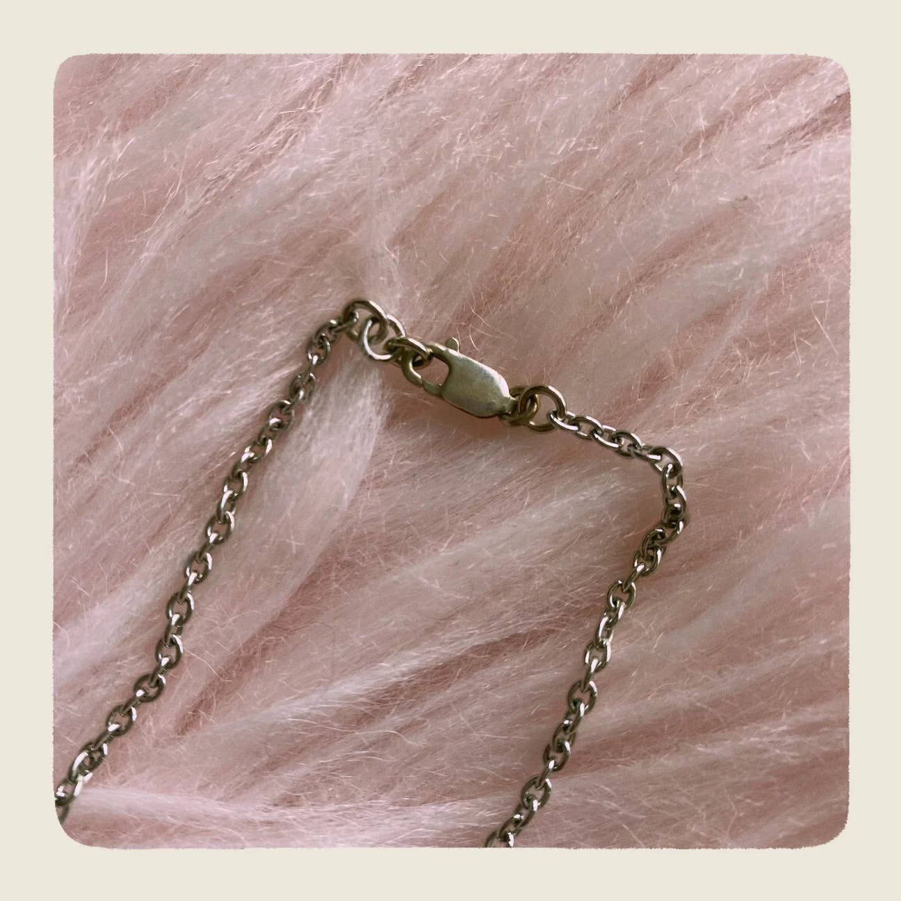 A close-up of a silver-toned metal chain necklace with a lobster clasp, laid on a soft pink textured faux-fur surface.