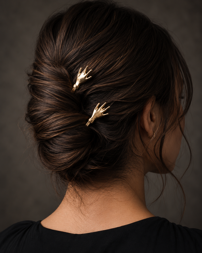 A woman shown from the back wearing a dark brown updo hairstyle with two small gold claw-shaped hair pins, against a dark gray studio background.