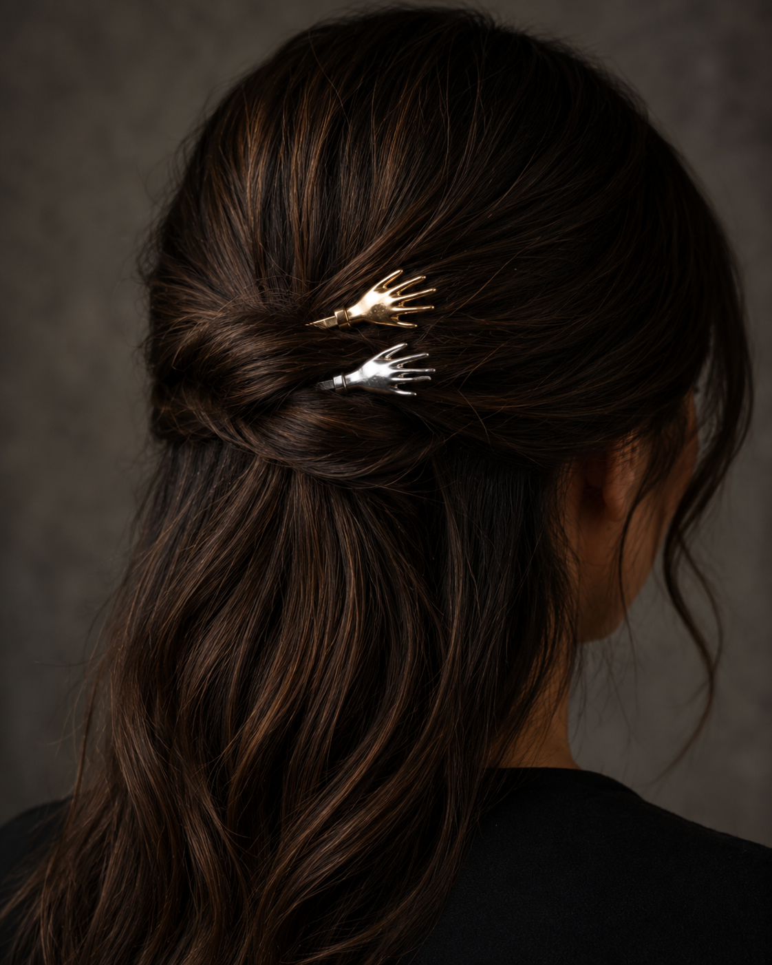 A close-up back view of a woman with long dark brown hair wearing two small skeleton hand hair clips in gold and silver, tucked into her low bun.