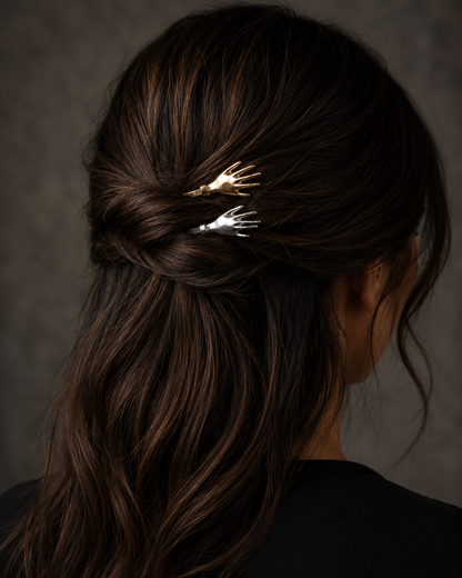 A close-up back view of a woman with long dark brown hair wearing two small skeleton hand hair clips in gold and silver, tucked into her low bun.
