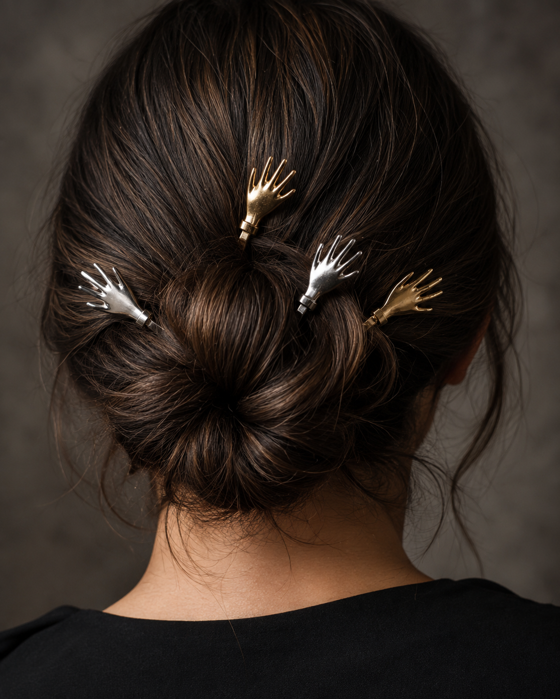 A person viewed from the back with dark brown hair styled in a low bun, wearing four small hand-shaped hair clips in silver and gold against a dark gray studio background.
