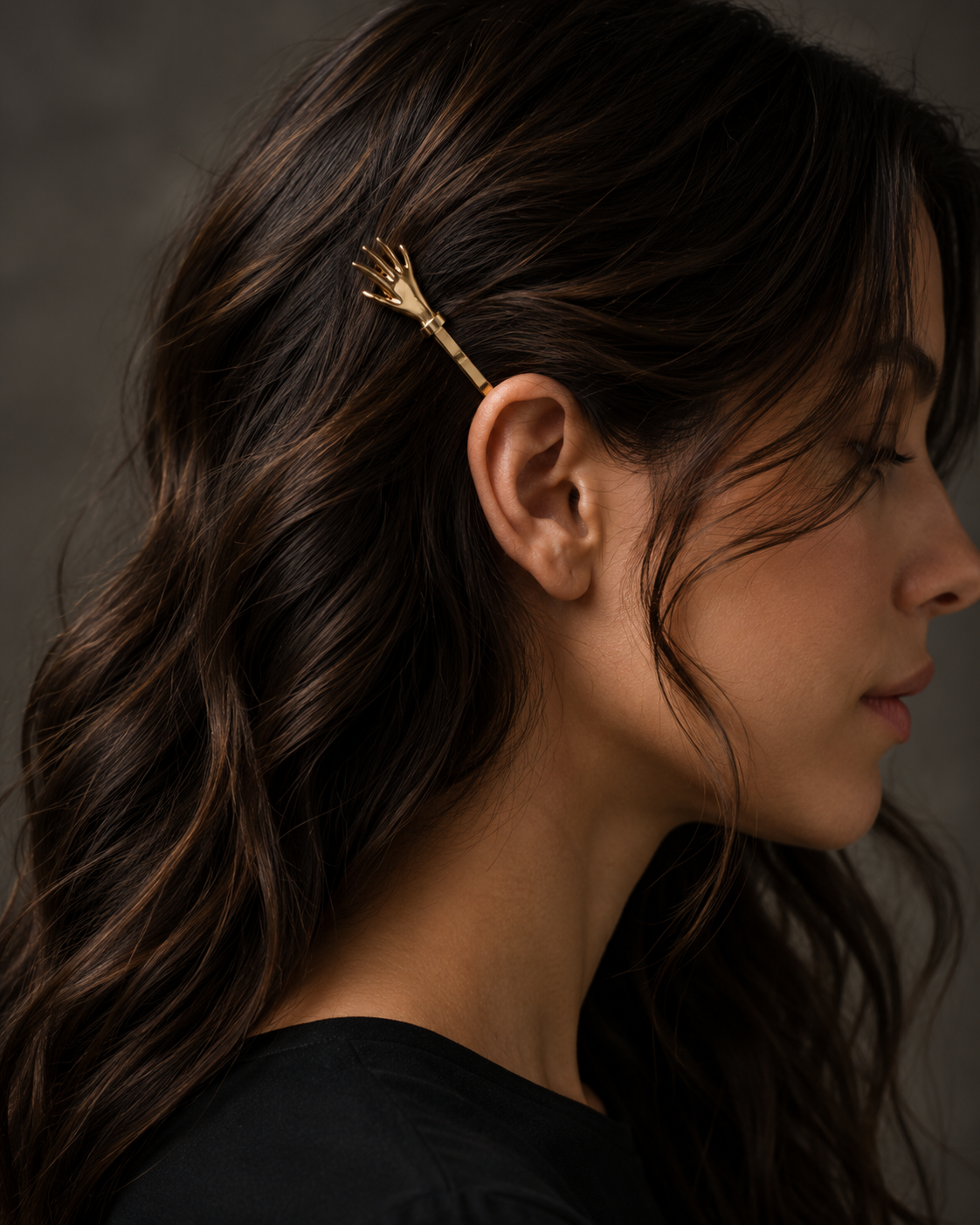 A close-up side profile of a woman with long, wavy dark brown hair wearing a small gold skeleton hand hair clip on the side of her head, set against a dark textured gray background.