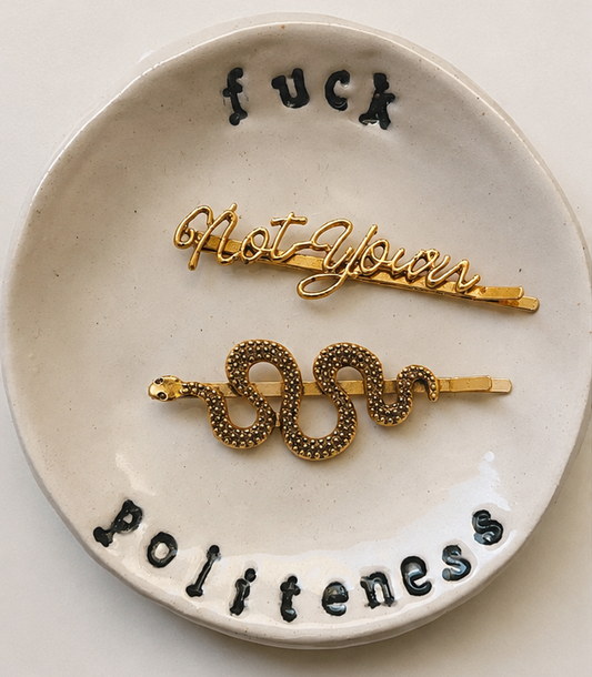 Two gold-tone metal hair claw clips displayed on a small round off-white ceramic dish. The top clip is a cursive script clip reading 'Not Yours', and the bottom clip is a coiled snake design with the word 'SAS' in raised gold letters.