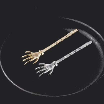 Two miniature skeleton hand hair pins, one gold-plated and one silver-plated, displayed on a dark circular surface.