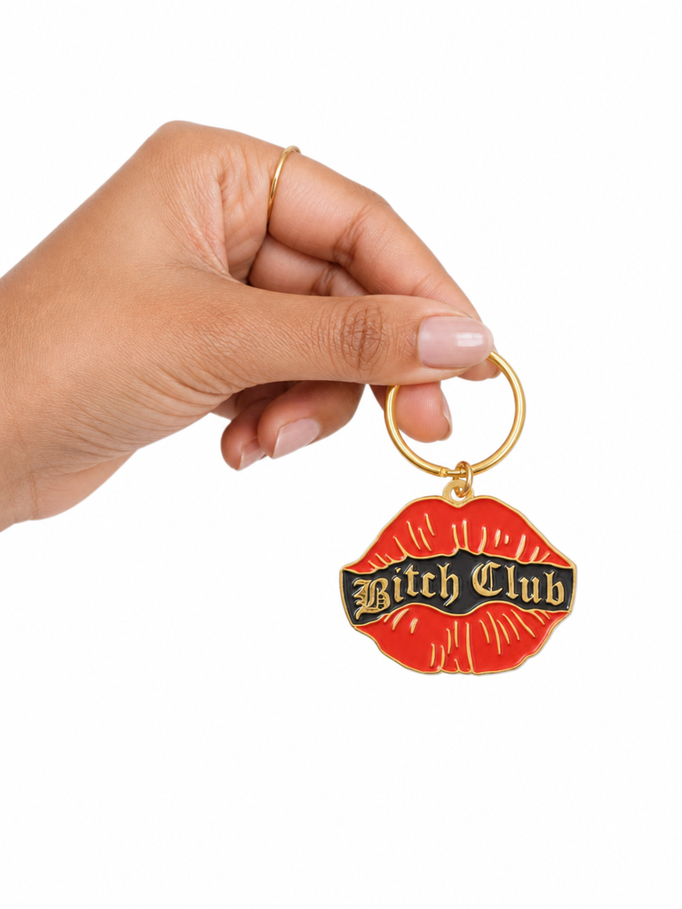 A hand holding a gold-tone keychain with a red lips-shaped charm featuring the text 'Bitch Club' in black gothic lettering on a white background.