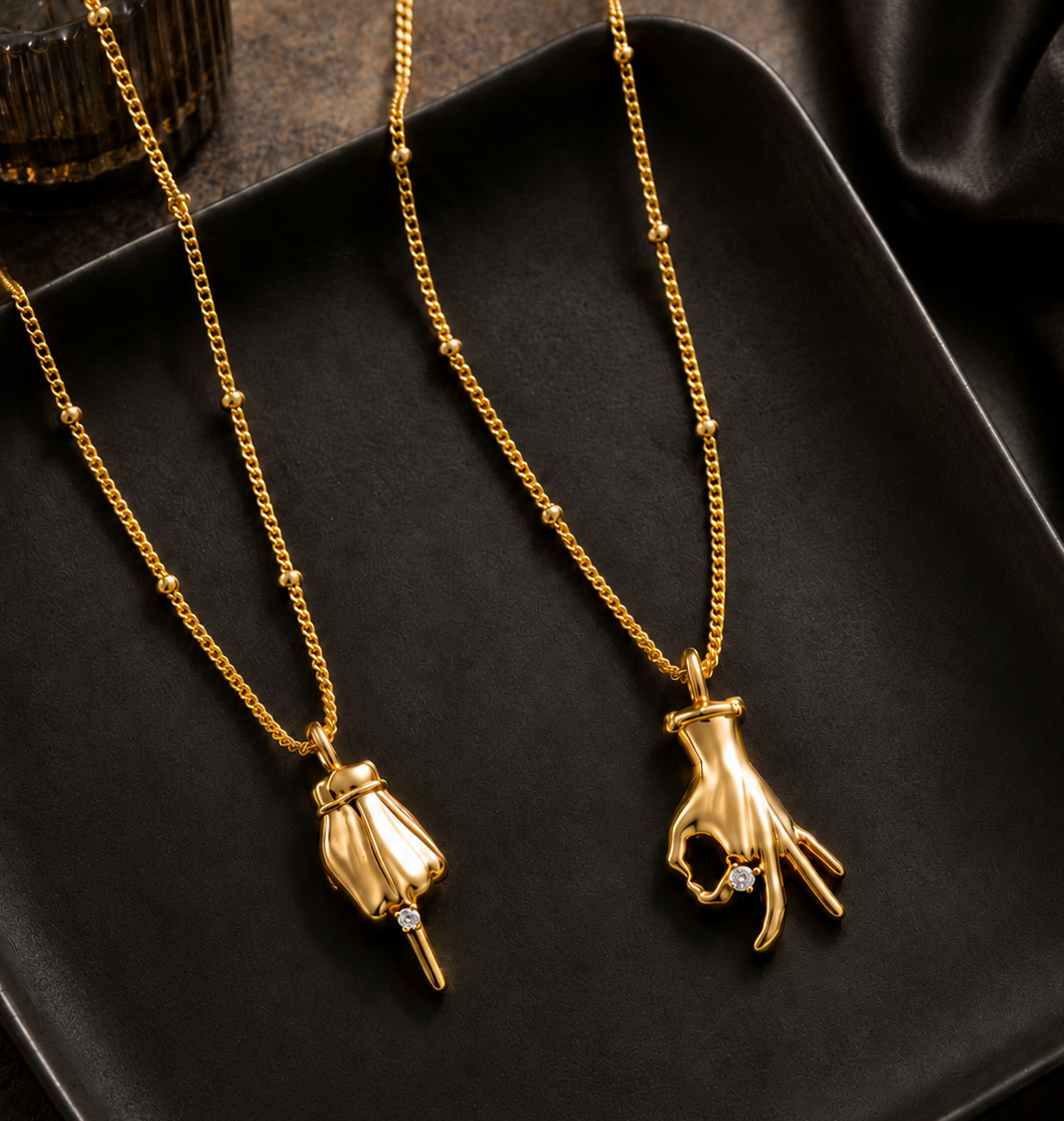 Two gold necklaces with unique pendant designs displayed on a black tray. One pendant is a textured gold bell with a small clear gem, and the other is a gold hand pendant holding a gold ring with a clear gem.