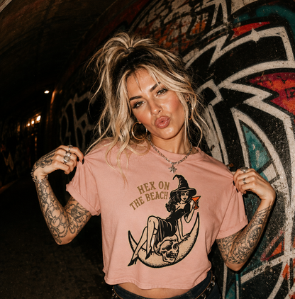 A woman with blonde hair in a messy bun poses in a graffiti-covered tunnel wearing a light pink graphic t-shirt that reads 'HEX ON THE BEACH' with a witch and skull illustration, paired with dark jeans.