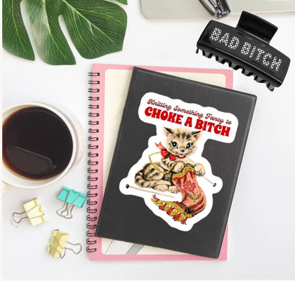 A black spiral notebook with a die-cut sticker featuring a kitten knitting while holding a pink bird, placed on top of a pink notebook. The notebook is styled on a white desk with a coffee cup, paper clips, a leaf, and a black hair clip that reads 'BAD BITCH'.