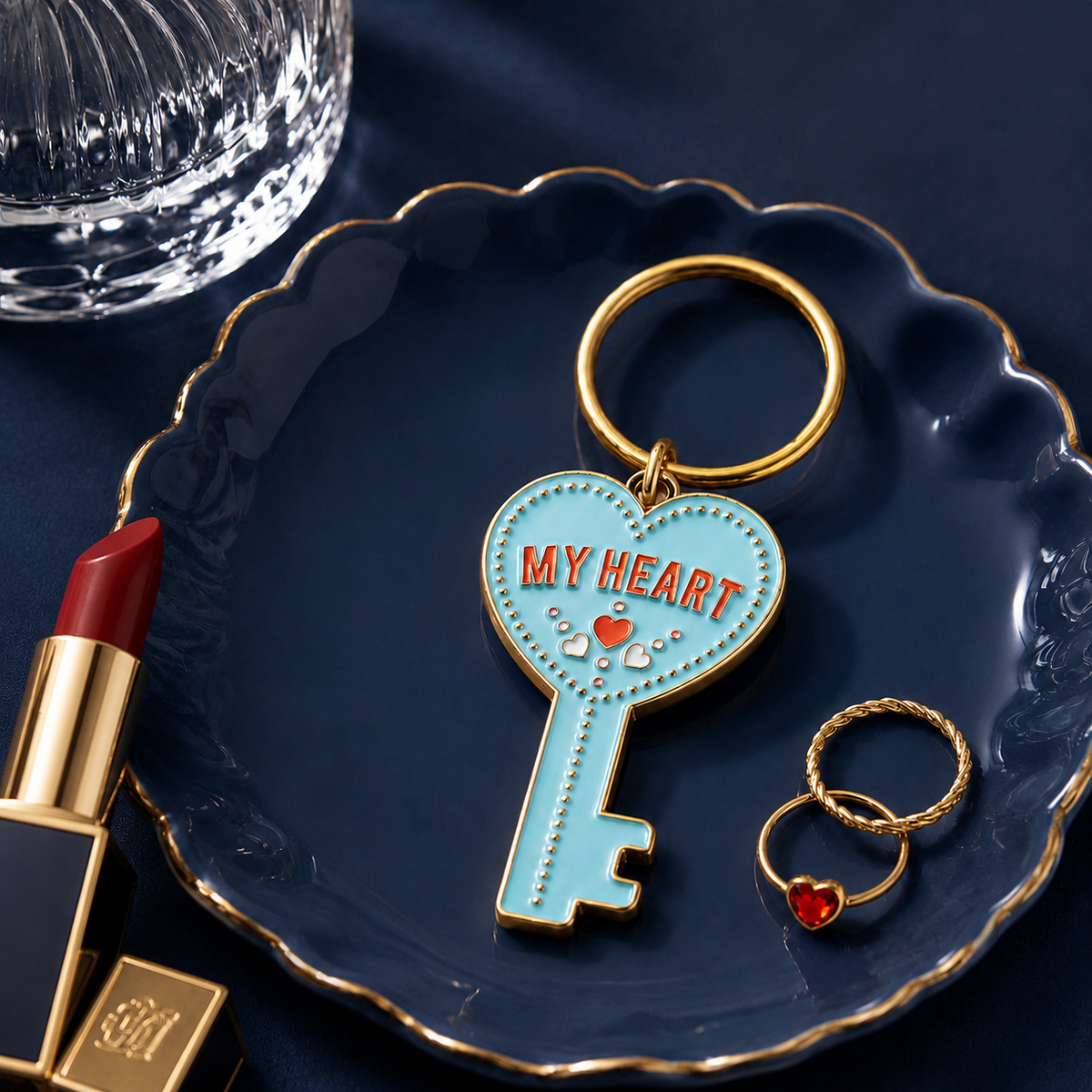 A light blue enamel key-shaped keychain with gold-tone metal hardware, featuring the words 'MY HEART' in red and small heart details, displayed on a dark blue scalloped plate with lipstick and rings nearby.