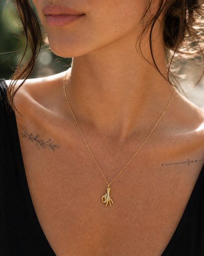 A close-up lifestyle shot of a woman wearing a delicate gold chain necklace with a small abstract gold pendant, set against a warm, sunlit outdoor background.