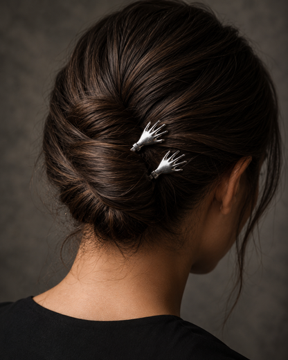 A close-up back view of a person with dark brown hair styled in a low bun, wearing two small silver hand-shaped hair pins. The background is a textured dark gray studio backdrop.