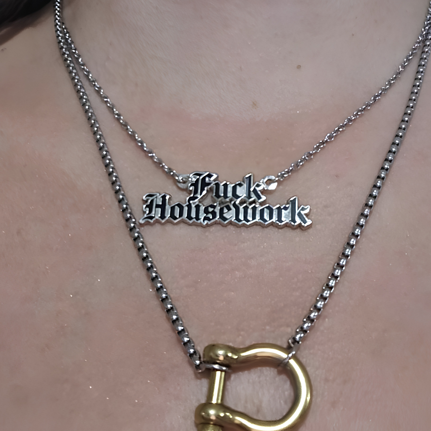 A close-up of a person's neck wearing two layered necklaces. The shorter silver chain features a gothic-style metal pendant that reads 'Fuck Housework' in black lettering.