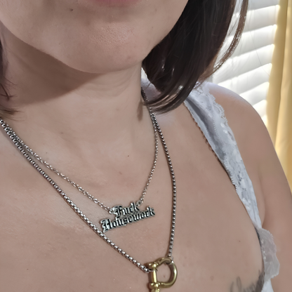 A close-up of a person wearing two layered silver necklaces, one with a gothic-style 'Fuck Housework' pendant and the other with a gold-colored D-ring clasp, paired with a white lace-trimmed camisole.