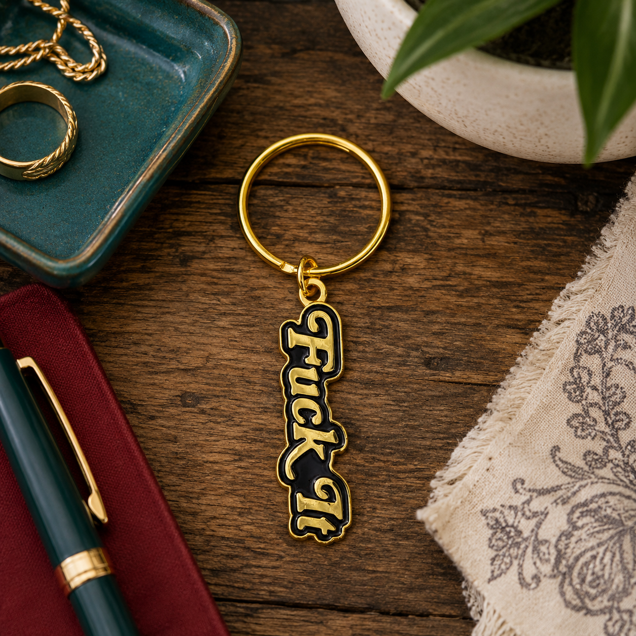 A gold-tone metal keychain with a vertical enamel charm spelling 'Fuck It' in black and white, displayed on a dark wood tabletop with lifestyle props including a pen, jewelry, and fabric.