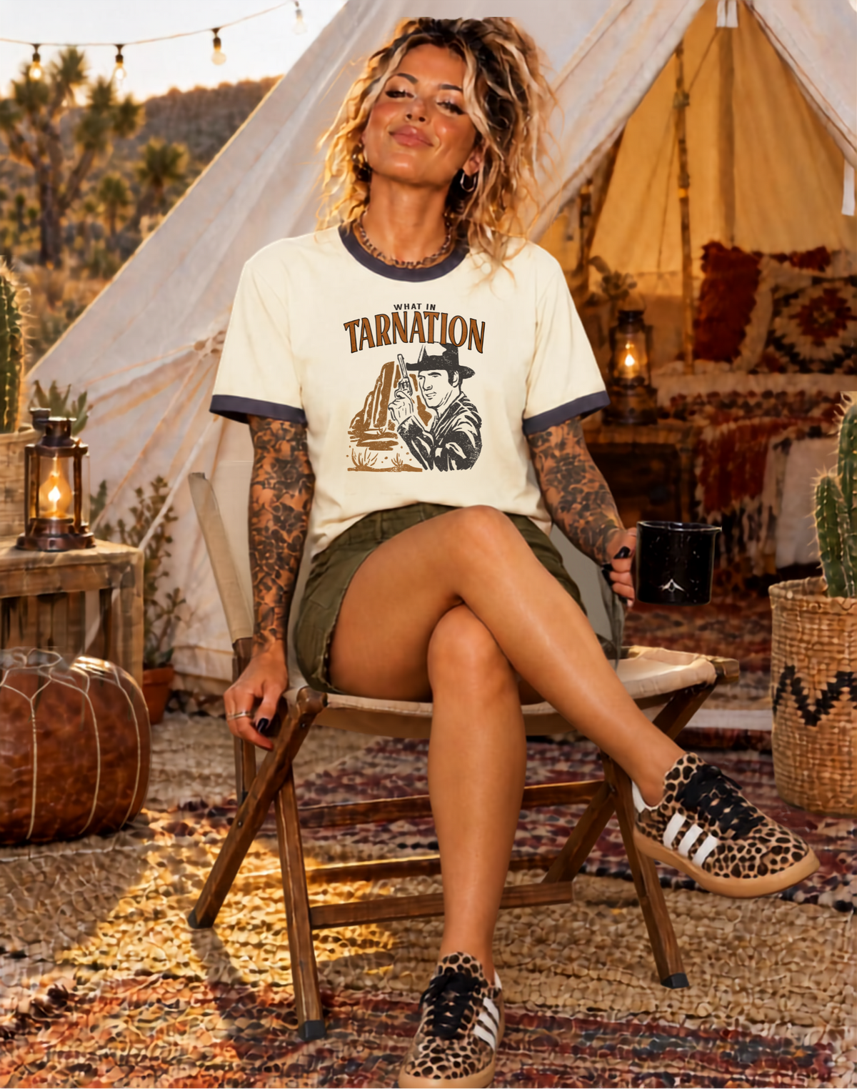A woman sits on a folding chair in a boho-style campsite setting, wearing a cream graphic t-shirt with dark blue trim and the word 'TARNATION' printed on the front, paired with olive green shorts and leopard-print sneakers.