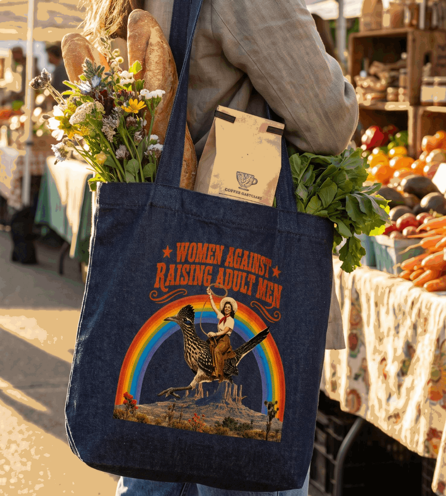 A woman at an outdoor farmers market carries a dark blue tote bag with a colorful graphic of a woman riding a horse in front of a rainbow and the text 'WOMEN AGAINST RAISING ADULT MEN'. The bag also contains bread, flowers, and a paper bag.