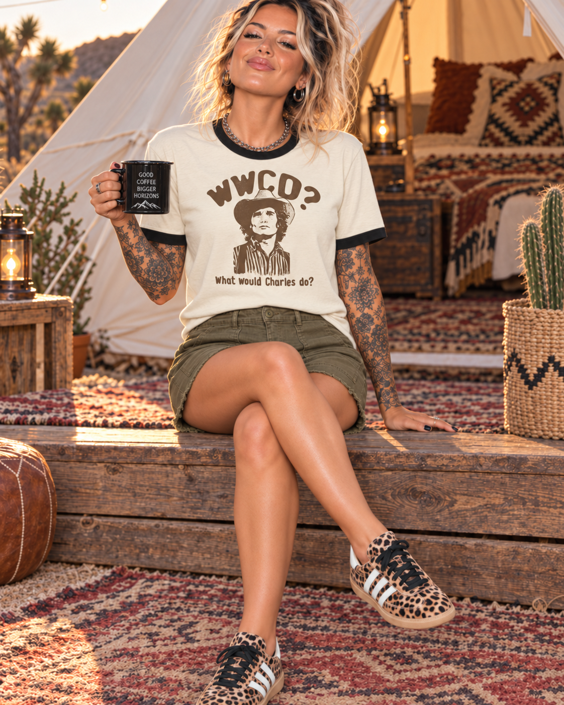 A woman sits on a rustic wooden bench in a desert-themed campsite setting, wearing a cream ringer t-shirt with a brown graphic print that reads 'WWCO? What would Charles do?'. She is also wearing olive green shorts and leopard print sneakers.